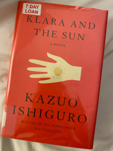 Book, Klara and the Sun, a novel by Kazuo Ishiguro. Hardcover book with red book sleeve, and white hand and a sun on its palm. 